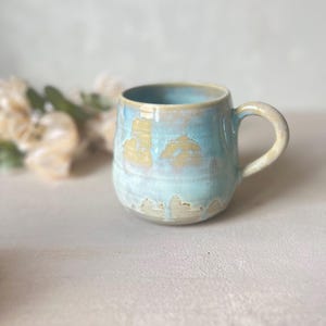 Peut inclure: Une tasse en céramique avec un corps arrondi et une anse incurvée. L'extérieur présente un émail réactif avec un mélange de tons bleu clair et beige sable, créant un effet superposé où le bleu coule sur la base beige vers le bas.