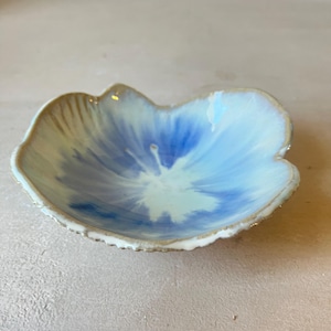 Op de afbeelding: Een kleine, ondiepe keramische kom met een onregelmatige, organische vorm die doet denken aan een bloemblad. De binnenzijde heeft een glanzend glazuur met een verloop van wit naar felblauw, terwijl de buitenrand een getextureerde, aardse afwerking heeft.