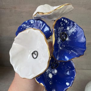 Peut inclure: Un groupe de cinq ornements en céramique en forme de fleurs aux bords ondulés. Trois fleurs sont bleu profond avec des motifs tachetés blancs, et deux sont blanc uni. Chaque fleur présente un bord peint en or et une petite boucle en métal sombre.