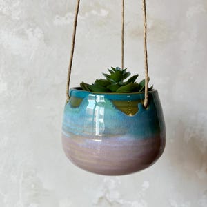 Puede incluir: Una maceta colgante de cerámica redonda con un esmalte brillante que presenta un degradado de azul verdoso intenso en la parte superior, que cambia a un suave rosa lavanda en la base. Una planta suculenta verde está dentro y cuelga de yute.