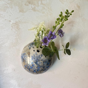 Peut inclure: Un vase mural rond en céramique de type pique-fleurs, doté d'une base crème tachetée ornée d'un motif floral bleu. Le vase présente plusieurs ouvertures circulaires sur le dessus, contenant un petit arrangement de fleurs artificielles violettes.