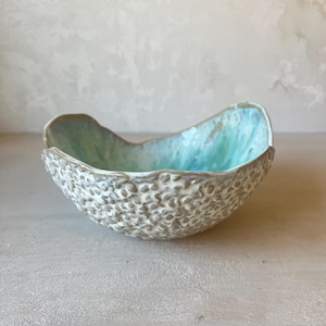 Peut inclure: Un bol en céramique avec un bord irrégulier et ondulé. L'extérieur présente une surface texturée et granuleuse dans des tons beige et blanc. L'intérieur est recouvert d'un émail bleu turquoise lisse et brillant, dégradé vers un vert pâle.