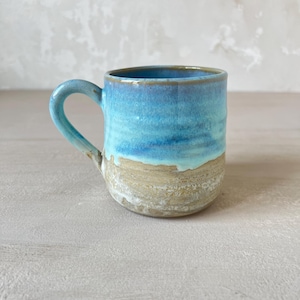 Peut inclure: Une tasse en céramique présentant un design bicolore avec un émail bleu vif et brillant sur la partie supérieure et une finition beige terreuse et texturée sur la moitié inférieure. La tasse comprend une anse incurvée et un bord arrondi.