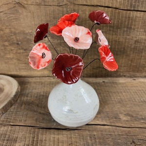 May include: A ceramic vase with a bouquet of red and pink poppy flowers. The vase is white with a speckled finish. The flowers have dark red centers and are arranged with thin, black stems.
