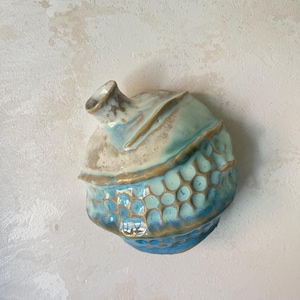 Op de afbeelding: Een keramische wandvaas met een ronde, asymmetrische vorm en een glanzend glazuur in blauw-, crème- en bruintinten. Het onderste gedeelte heeft een verhoogde, honingraatachtige textuur, terwijl het bovenste deel glad is met een kleine opening.