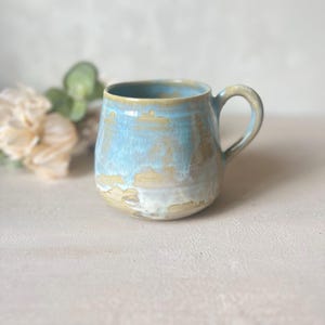 Può includere: Una tazza in ceramica con corpo arrotondato e manico curvo, caratterizzata da una finitura a smalto reattivo che fonde tonalità di azzurro, crema e marrone terroso in un motivo a strati e gocciolante sulla superficie esterna e sul bordo.