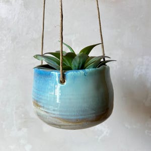 Op de afbeelding: Een ronde hangende keramische plantenpot met een glanzend glazuur dat verloopt van een levendig hemelsblauw aan de bovenkant naar een gedempt aards bruin aan de basis. De pot hangt aan een dun, natuurkleurig touw en bevat een groene vetplant.