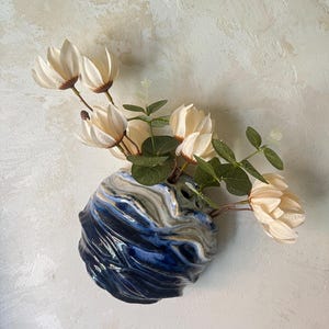 Peut inclure: Un vase en céramique fixé au mur présentant un motif marbré tourbillonnant de bleu marine profond, de blanc et de brun clair. Le récipient arrondi et texturé contient un bouquet de fleurs de magnolia crème artificielles et de feuilles d'eucalyptus vertes.