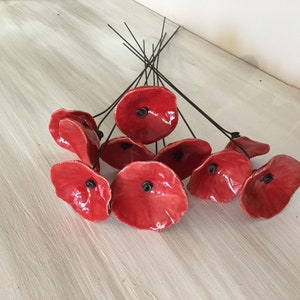 Pode incluir: Um grupo de papoilas de cerâmica feitas à mão em um tom vermelho vibrante. Cada flor apresenta um centro escuro e é presa a um caule de arame preto. As papoilas estão dispostas sobre uma superfície clara.