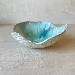 Peut inclure: Un petit bol en céramique à la forme irrégulière et organique. L'intérieur présente une glaçure brillante dans des tons turquoise, bleu et blanc, tandis que l'extérieur a une finition mate blanc cassé texturée avec des stries horizontales.