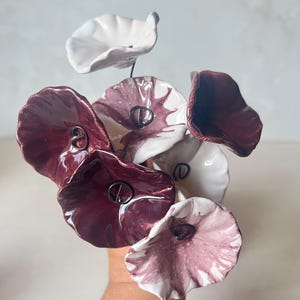 Könnte beinhalten: Ein Strauß aus sechs handgefertigten Keramikblumen mit gewellten, organisch geformten Blütenblättern. Die Blumen haben eine glänzende Oberfläche in den Farben Dunkelburgunder, zartes Rosa und Weiß. Jede Blütenmitte ist mit einem dunklen Drahtakzent versehen.