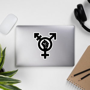 May include: Black and white sticker featuring a transgender symbol with a raised fist in the centre.
