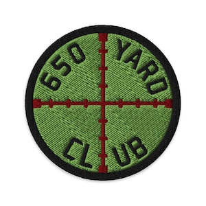 May include: Embroidered patch with a green background and a red crosshair design. The text "650 YARD CLUB" is stitched in black around the perimeter. The patch has a black border.