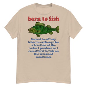 Puede incluir: Camiseta color beige con la frase "born to fish" en rojo y una ilustración de un pez verde. Debajo, texto azul: "forced to sell my labor in exchange for a fraction of the value i produce so i can afford to fish on the weekend sometimes."