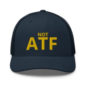 Puede incluir: Una gorra de camionero azul marino con una malla negra en la parte posterior. La gorra tiene las palabras "NOT ATF" bordadas en amarillo en la parte delantera.