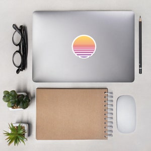 May include: A laptop computer with a sticker of a sunset with a retro color scheme. The sticker is a circle with a yellow, orange, and purple gradient. The laptop is on a white surface with a notebook, a mouse, and two small potted plants.