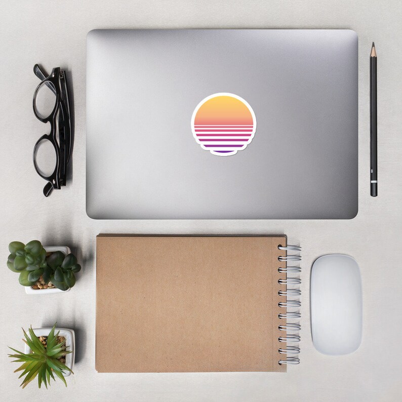 May include: A laptop computer with a sticker of a sunset with a retro color scheme. The sticker is a circle with a yellow, orange, and purple gradient. The laptop is on a white surface with a notebook, a mouse, and two small potted plants.