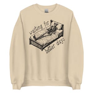Może przedstawiać: Beżowa bluza z nadrukiem szkieletu leżącego w łóżku i tekstem "waiting for better days".