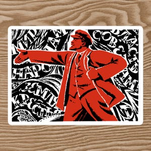 Può includere: Un adesivo rettangolare con un'illustrazione stilizzata di un uomo in abito e berretto rossi, che indica con il braccio sinistro. Lo sfondo è bianco e nero con testo astratto. L'adesivo è su una superficie di legno.