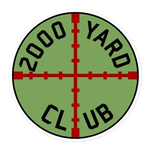 Puede incluir: Una pegatina circular verde con un diseño de diana de retícula roja y el texto "2000 YARD CLUB" en negro.