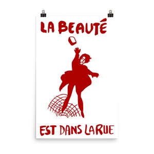 May include: A red silhouette of a person throwing a brick at a fence with the text "LA BEAUTÉ EST DANS LA RUE" in red.