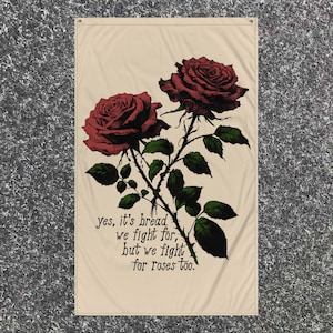 Puede incluir: Una bandera de lona beige con dos rosas rojas y hojas verdes. La bandera tiene el texto "yes, it's bread we fight for, but we fight for roses too."