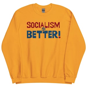 May include: Mustard yellow sweatshirt featuring the slogan "SOCIALISM IS BETTER!" in red and blue lettering. The crew neck sweatshirt has long sleeves. The text is centrally positioned on the front of the garment.