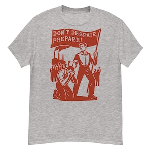 Op de afbeelding: Grijs t-shirt met een rode afbeelding. De afbeelding toont een man die een vlag vasthoudt met de tekst "DON'T DESPAIR, PREPARE!". Het bevat ook een knielende figuur en fabrieksschoorstenen.