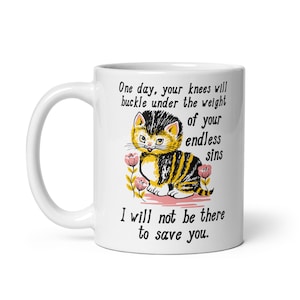 May include: White ceramic mug featuring a black and yellow cartoon cat with the text "One day, your knees will buckle under the weight of your endless sins I will not be there to save you."