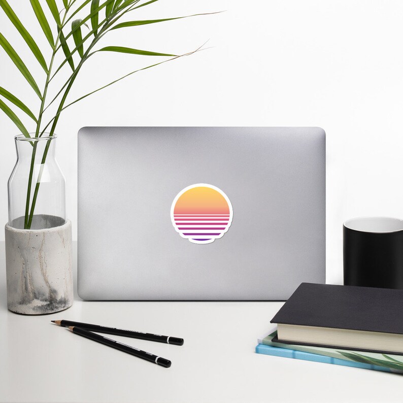 May include: A laptop computer with a sticker of a retro sunset design. The sticker is a circle with a yellow, orange, and pink gradient and horizontal lines in purple and white.