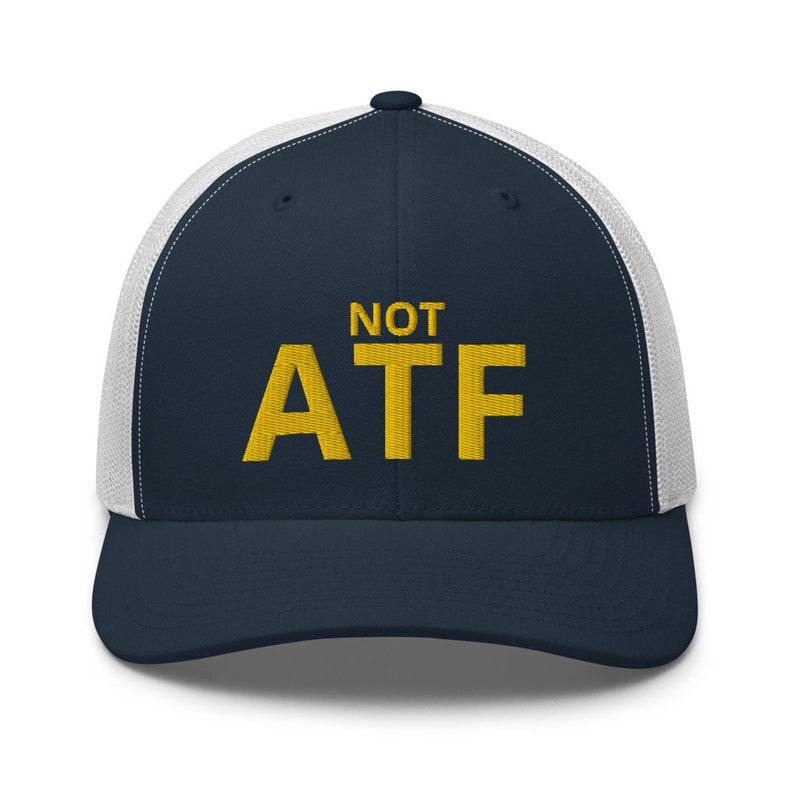 Puede incluir: Una gorra de camionero azul marino y blanca con el texto "NOT ATF" bordado en amarillo.
