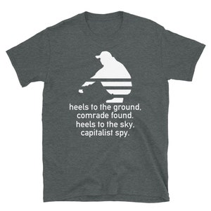 Pode incluir: Uma camiseta cinza escura mesclada com uma silhueta branca de uma pessoa sentada com o texto "heels to the ground, comrade found. heels to the sky, capitalist spy."