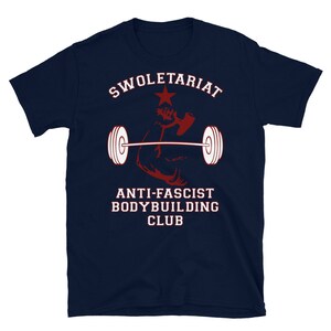 May include: A navy blue t-shirt with the text "Swole-tariat Anti-Fascist Bodybuilding Club" in white and red lettering. A red star and a red silhouette of a bicep lifting a barbell are also featured on the shirt.