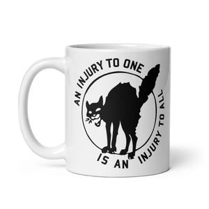May include: White ceramic mug with a black illustration of a cat in a circle. The text around the circle reads "AN INJURY TO ONE IS AN INJURY TO ALL". The mug has a curved handle.