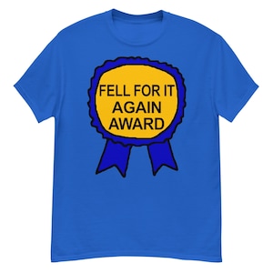 Puede incluir: Camiseta azul con un diseño de cinta de premio amarilla con el texto negro "FELL FOR IT AGAIN AWARD". La cinta tiene un borde azul y dos cintas azules.