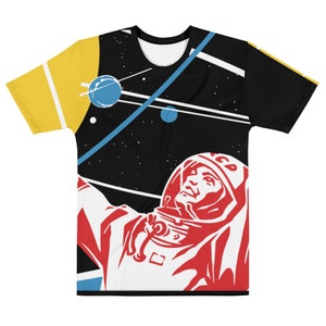May include: A short-sleeved t-shirt featuring a retro space-themed design. The shirt has a black background with a graphic of an astronaut in a red spacesuit, blue planets, and white lines. The sleeves are yellow and black.