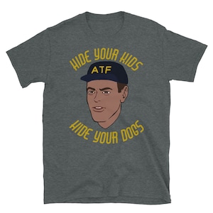 May include: A dark heather grey t-shirt with a cartoon illustration of a person wearing a blue baseball cap with the letters "ATF" on it. The text "Hide your kids, hide your dogs" is written in yellow around the illustration.