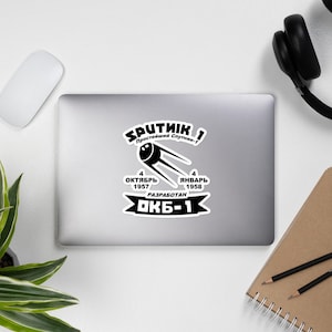 May include: Black and white sticker with a graphic of Sputnik 1, the first artificial satellite launched into space. The sticker includes the text "SPUTNIK 1", "Простейший Спутник-1", "4 ОКТЯБРЬ 1957", "4 ЯНВАРЬ 1958", "РАЗРАБОТАН", and "ОКБ-1".