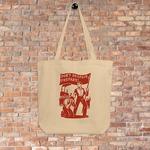 Op de afbeelding: Beige draagtas met een rode afbeelding. De afbeelding toont een man die een vlag vasthoudt met de tekst "DON'T DESPAIR, PREPARE!" en een menigte mensen. De tas heeft lange hengsels en hangt tegen een bakstenen muur.