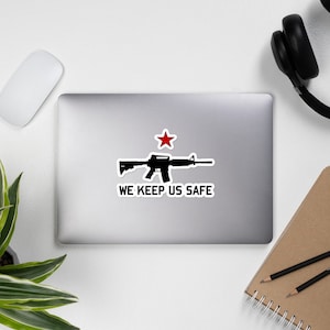 Può includere: Un adesivo bianco e nero con una silhouette di un fucile e una stella rossa sopra. Il testo "WE KEEP US SAFE" è sotto il fucile.