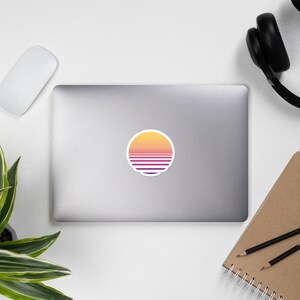 May include: A round sticker with a retro sunset design. The sun is yellow, orange, and pink with purple and white stripes.