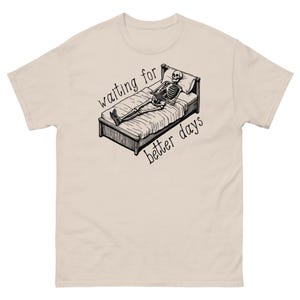 Może przedstawiać: Beżowa koszulka z czarno-białym rysunkiem szkieletu leżącego w łóżku. Na koszulce nadrukowany jest tekst "waiting for better days".