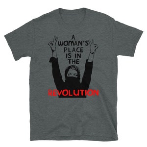 May include: A dark heather grey t-shirt with a black and white graphic of a woman with her arms raised in the air. The text "A Woman's Place Is In The Revolution" is printed in black on the shirt.