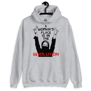 May include: A grey hooded sweatshirt with a black and white illustration of a person with their arms raised in the air. The text "A Woman's Place Is In The Revolution" is printed in black and red on the sweatshirt.