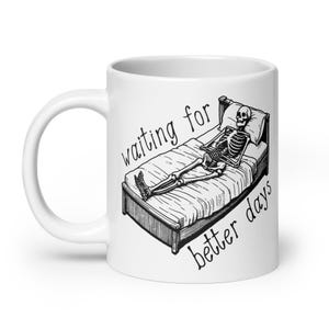 Może przedstawiać: Biały ceramiczny kubek z czarno-białą ilustracją szkieletu w łóżku. Na kubku znajduje się napis "waiting for better days" w stylu pisma odręcznego.