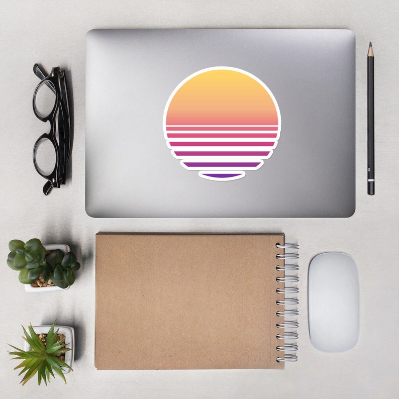 May include: A laptop computer with a sticker of a sunset with a retro color scheme. The sticker is a circle with a yellow, orange, pink, and purple color gradient. The sunset has five horizontal stripes in a purple and pink color scheme.