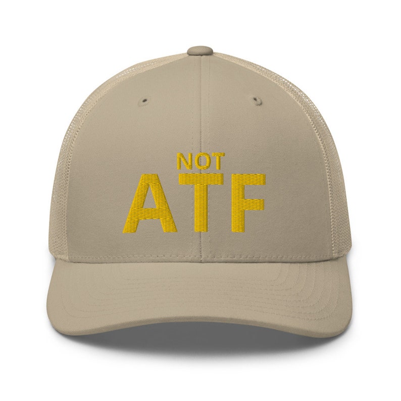 Puede incluir: Una gorra de camionero beige y negra con un dise&ntilde;o bordado en amarillo que dice "NOT ATF".
