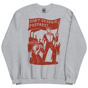 Op de afbeelding: Een grijze sweatshirt met een rode afbeelding. De afbeelding toont een man die een vlag vasthoudt met de tekst "DON'T DESPAIR, PREPARE!" en een knielende figuur. Het ontwerp bevat fabriekssilhouetten en andere figuren.
