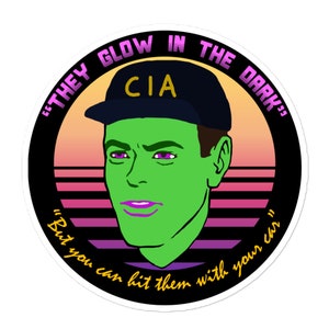 May include: A round sticker with a black background and a retro sunset design. The sticker features a cartoon illustration of a person wearing a black baseball cap with the letters "CIA" in white. The person has green skin and purple lips. The text around the image reads "They Glow in the Dark" and "But you can hit them with your car."