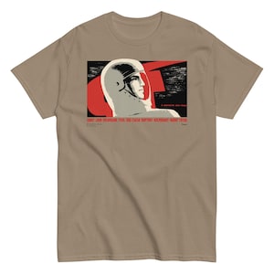 May include: A tan short-sleeve t-shirt featuring a graphic print of a cosmonaut in a helmet against a black and red background. The print includes Cyrillic text and a profile view of a person looking to the right, rendered in a vintage poster style.